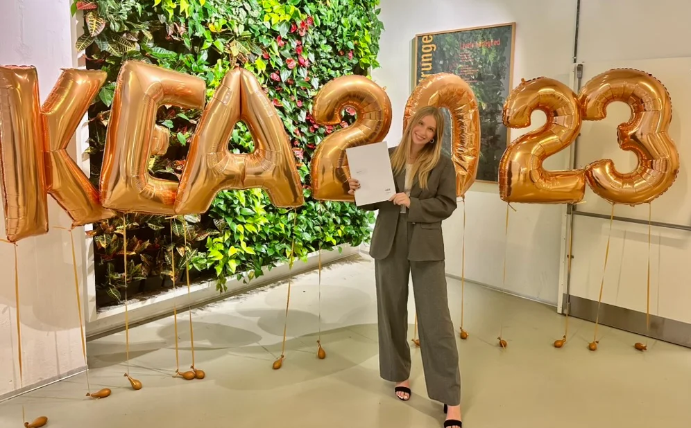 Młoda dziewczyna w zielonym garniturze i szpilkach stoi z dyplomem w dłoni. W tle duże balony z napisem KEA 2023.