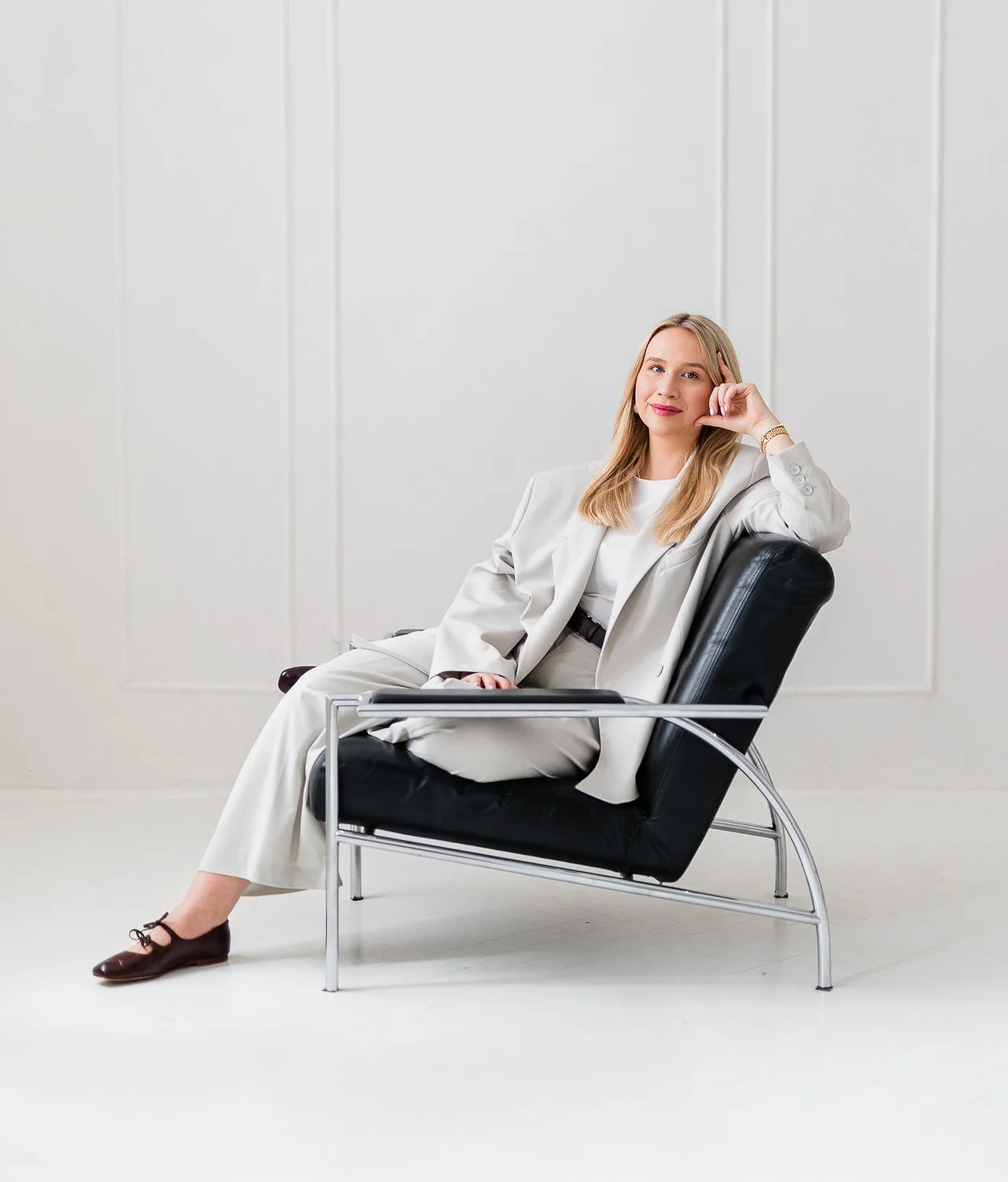 Młoda kobieta ubrana w stylowy biały garnitur siedzi na czarnym krześle i lekko uśmiecha się. Ma blond włosy.