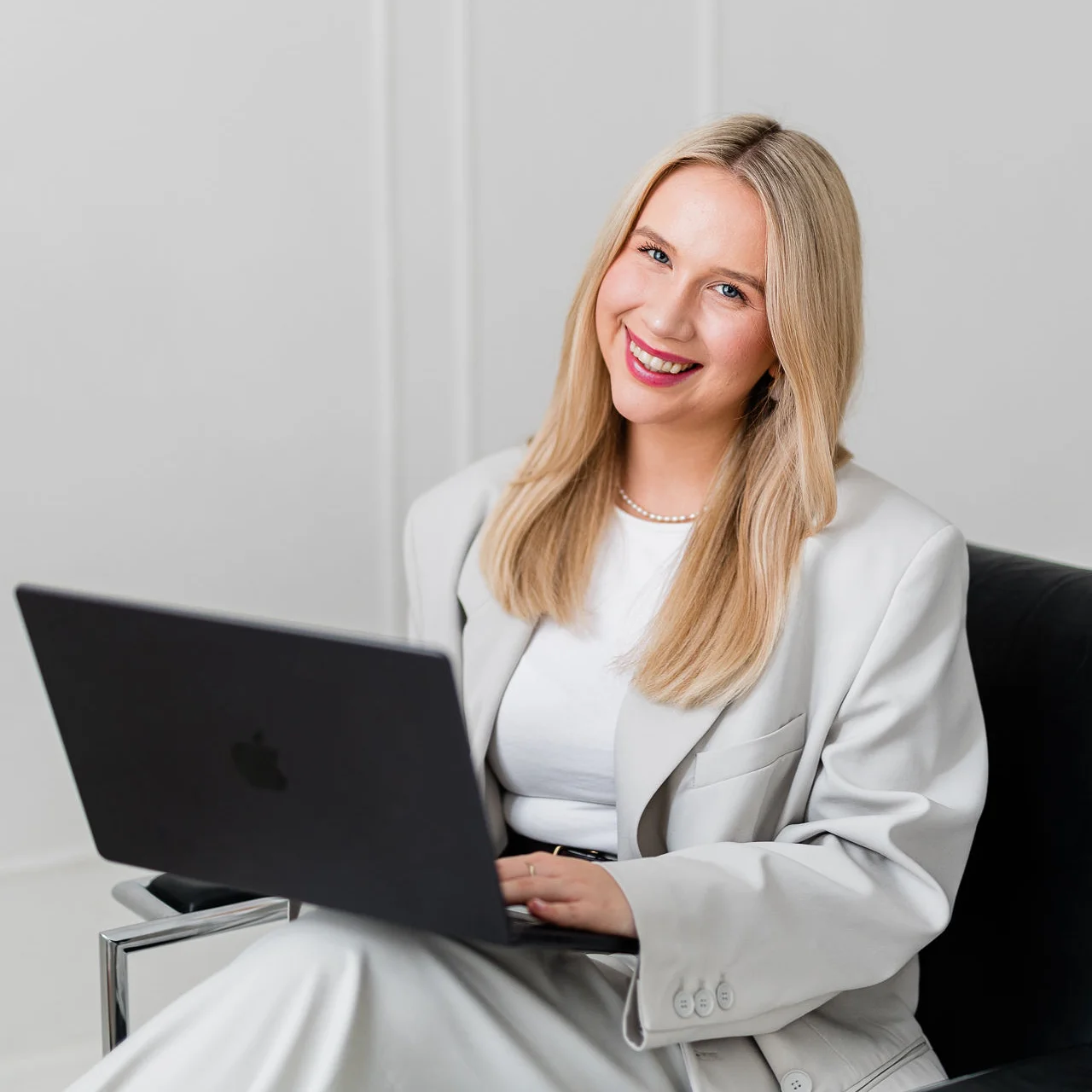 Młoda kobieta ubrana w stylowy biały garnitur siedzi na czarnym krześle i lekko uśmiecha się. Ma blond włosy i trzyma w dłoni laptopa.