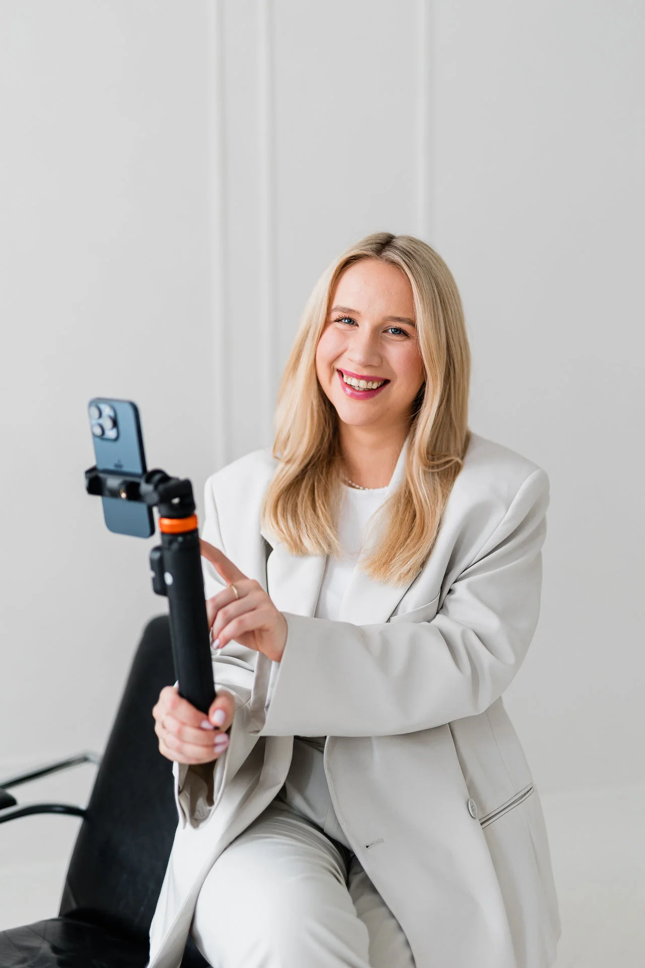 Młoda kobieta ubrana w stylowy biały garnitur siedzi na czarnym krześle i lekko uśmiecha się. Ma blond włosy i trzyma w statyw z telefonem.