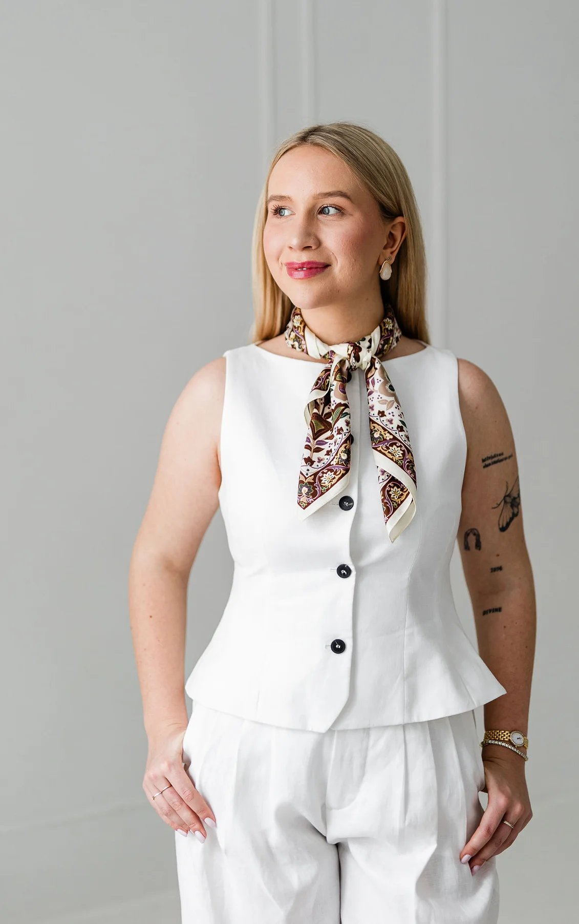 Młoda kobieta o blond włosach ubrana w białą kamizelkę i spodnie. Spogląda w swój prawy bok.