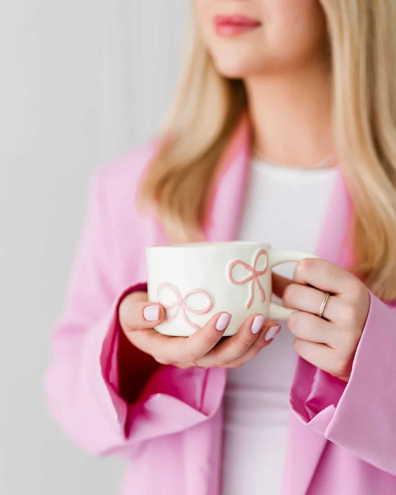 Młoda kobieta o blond włosach stoi z białą filiżanką. Ma na sobie różowy garnitur.