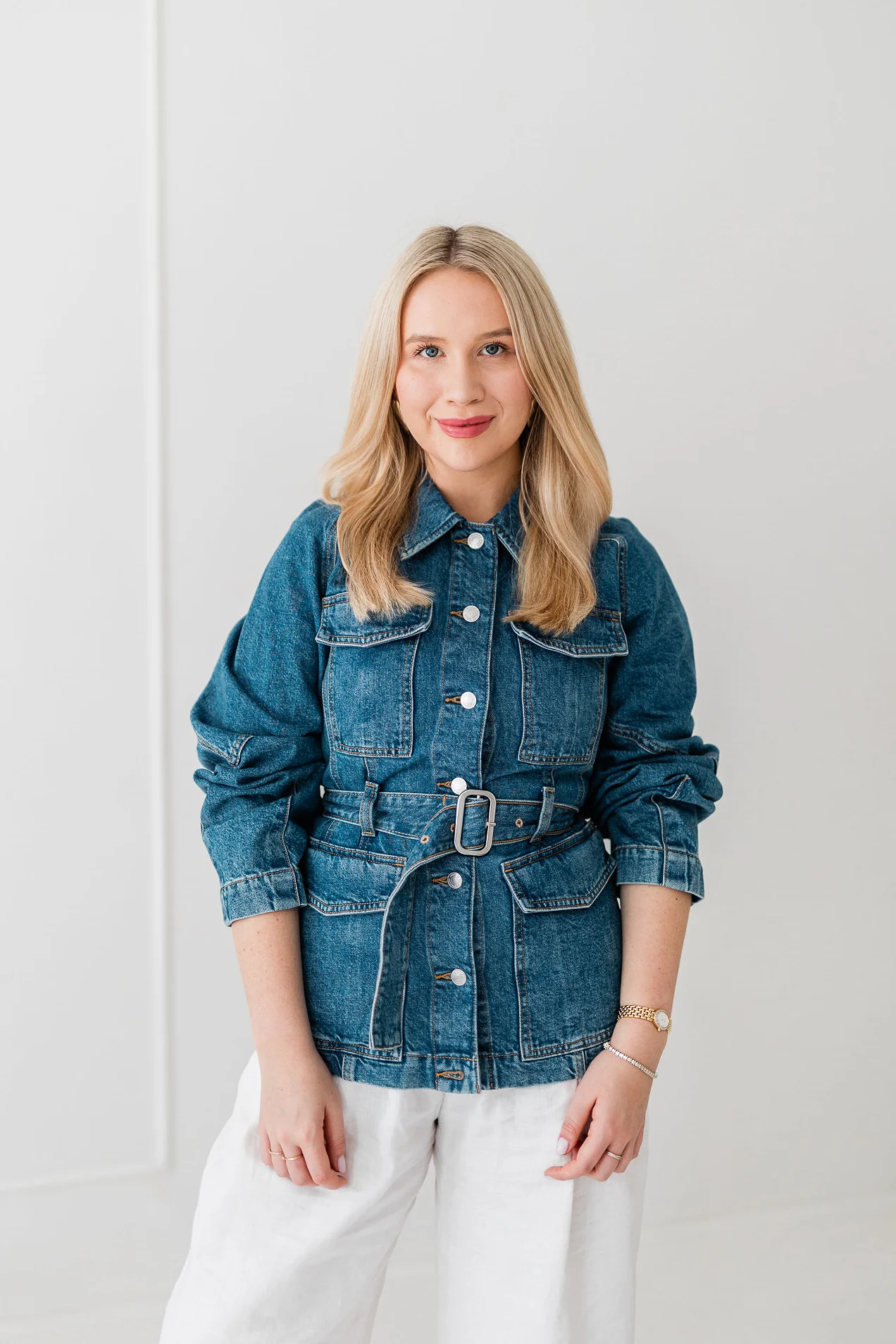 Młoda lekko uśmiechnieta kobieta o blond włosach stoi na białym tle. Ma na sobie złotą i srebrną widoczną biżuterię oraz dżinsowy żakiet.