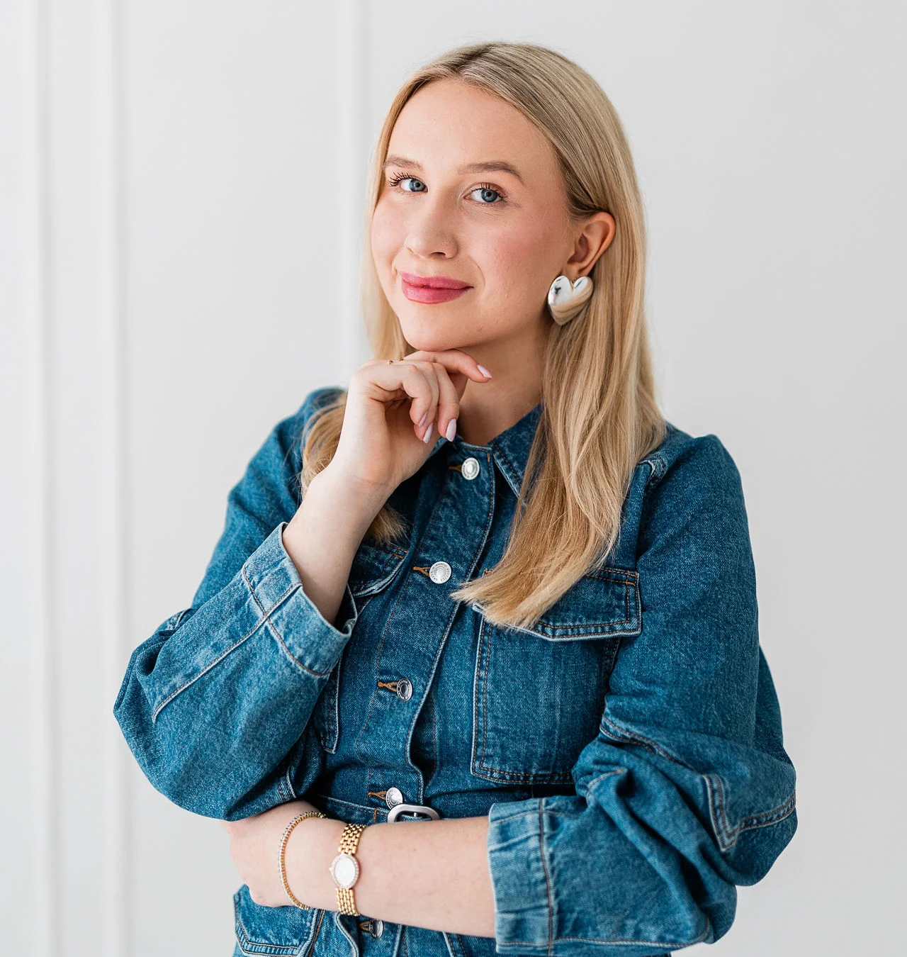 Młoda lekko uśmiechnieta kobieta o blond włosach stoi na białym tle. Ma na sobie złotą i srebrną widoczną biżuterię oraz dżinsowy żakiet.