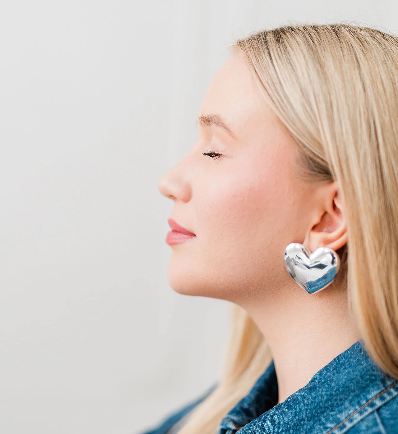 Lewy profil twarzy młodej kobiety o blond włosach. Na sobie ma duże srebrne kolczyki serca.