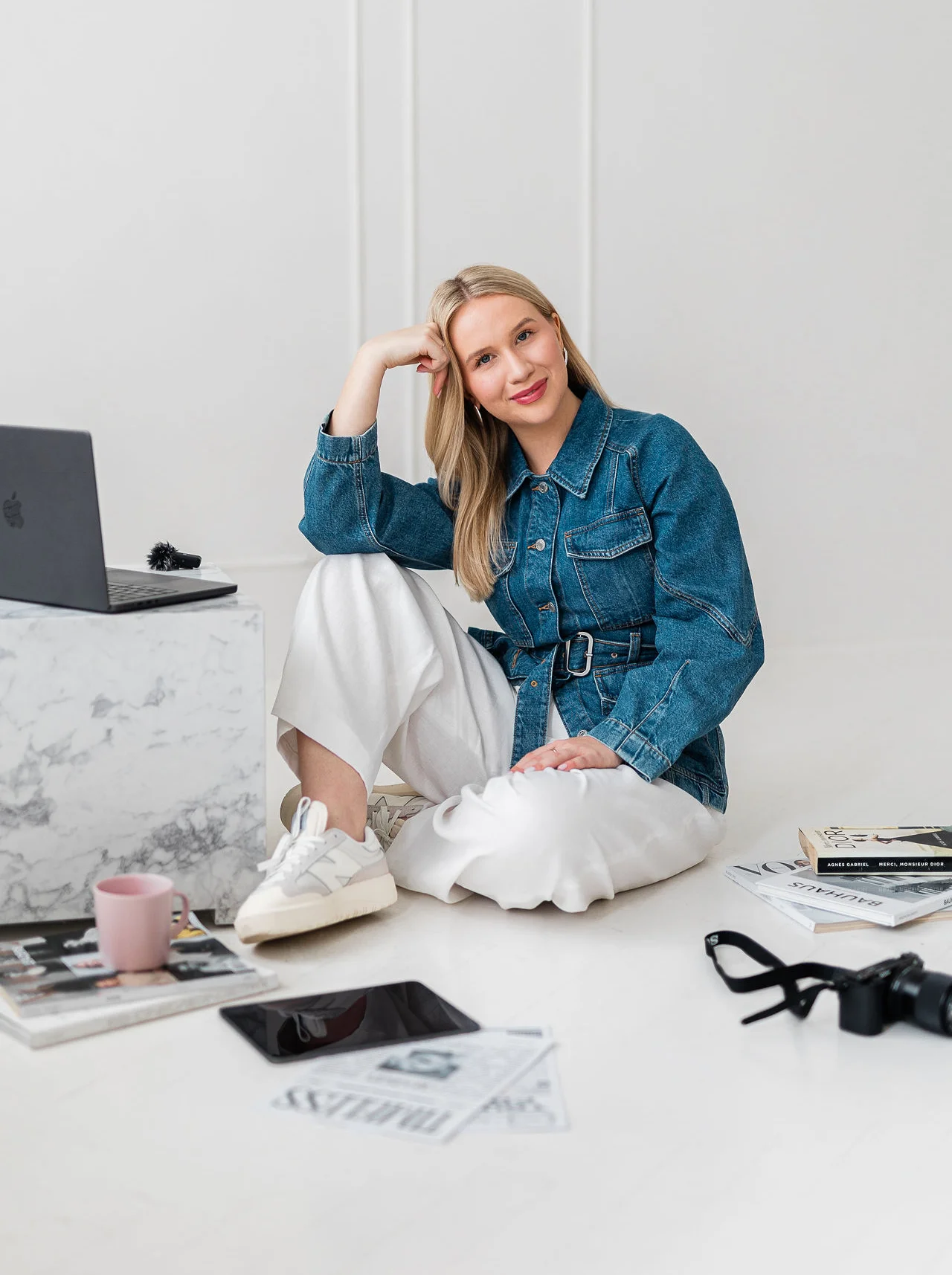 Młoda lekko uśmiechnięta kobieta o blond włosach siedzi na ziemi i korzysta z czarnego laptopa. Ma na sobie złotą i srebrną widoczną biżuterię oraz dżinsowy żakiet.