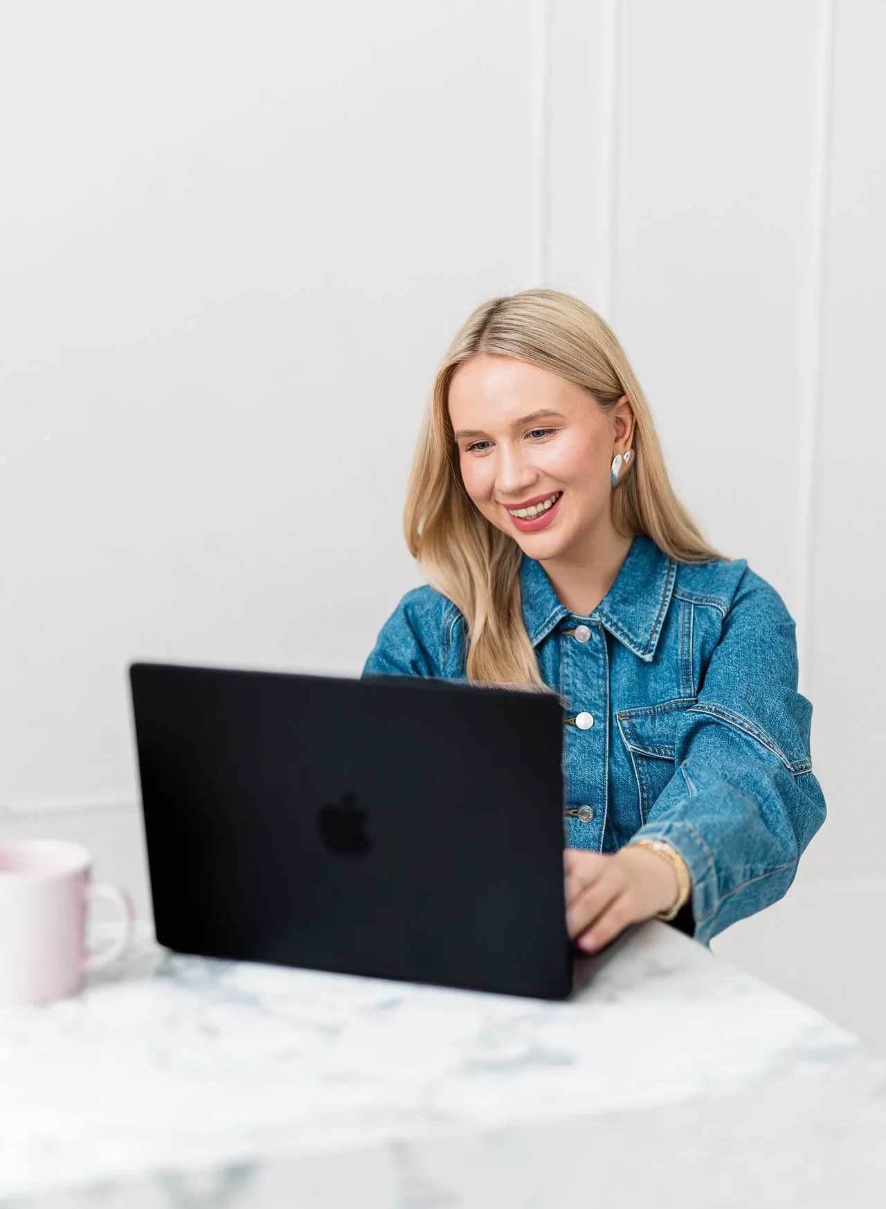 Młoda lekko uśmiechnieta kobieta o blond włosach siedzi przy biurku i korzysta z czarnego laptopa. Ma na sobie złotą i srebrną widoczną biżuterię oraz dżinsowy żakiet.