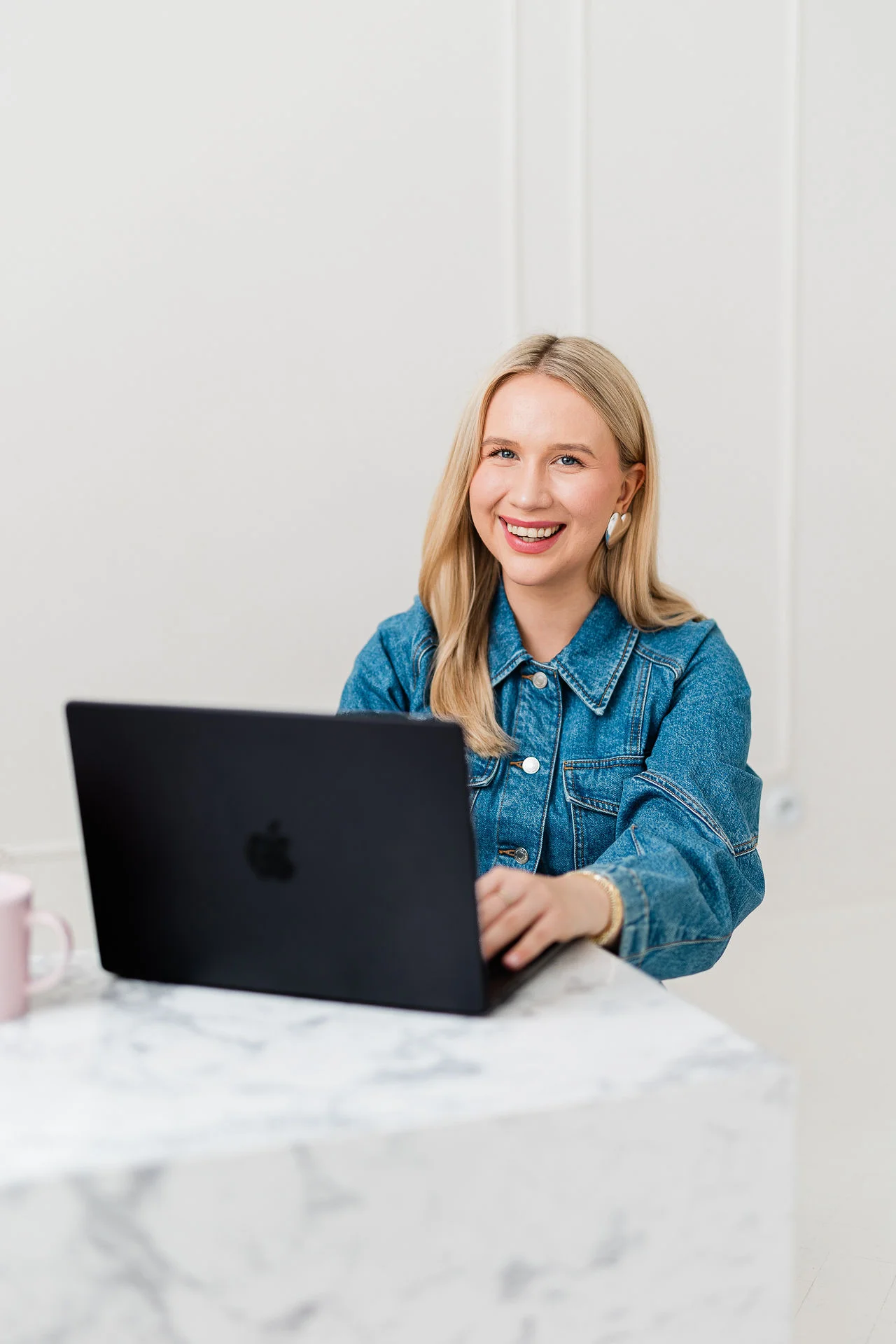 Młoda lekko uśmiechnieta kobieta o blond włosach siedzi przy biurku i korzysta z czarnego laptopa. Ma na sobie złotą i srebrną widoczną biżuterię oraz dżinsowy żakiet.