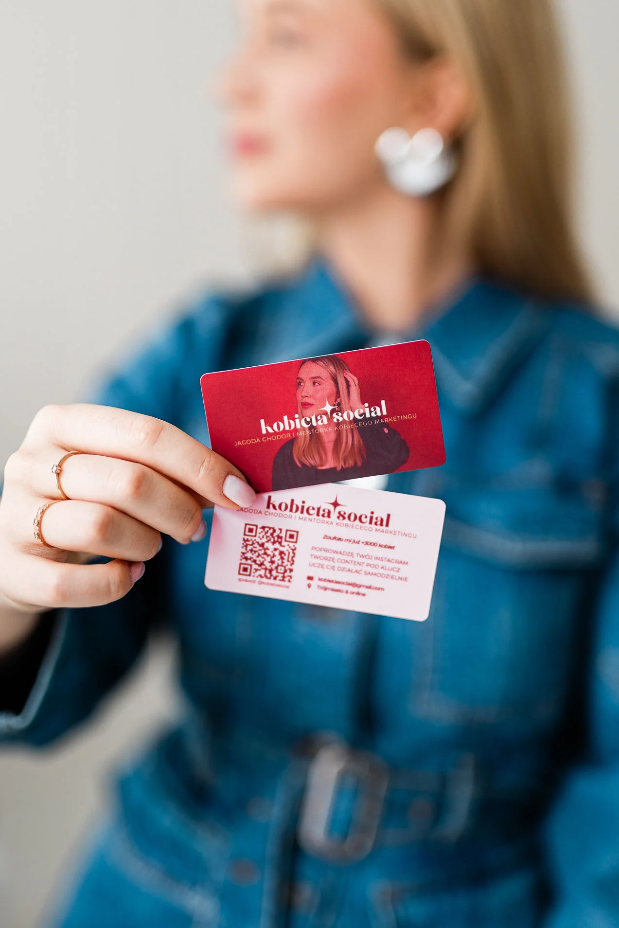 Młoda lekko uśmiechnieta kobieta o blond włosach stoi na białym tle. Ma na sobie złotą i srebrną widoczną biżuterię oraz dżinsowy żakiet. Przed sobą pokazuje dwie wizytówki.