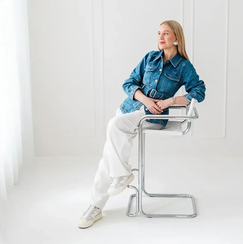 Młoda kobieta o blond włosach siedzi na wysokim metalowym krześle ubrana w dżinsowy żakiet i białe spodnie.