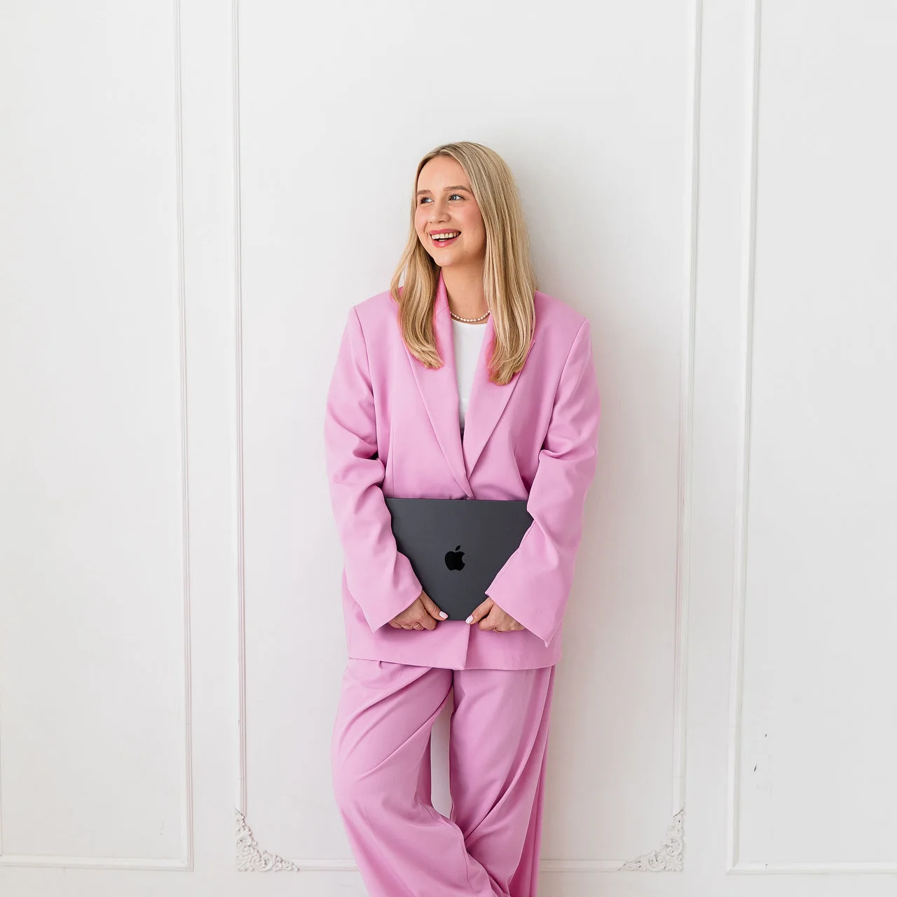 Młoda kobieta o blond włosach stoi z laptopem w dłoniach i patrzy w bok. Ma na sobie różowy garnitur.