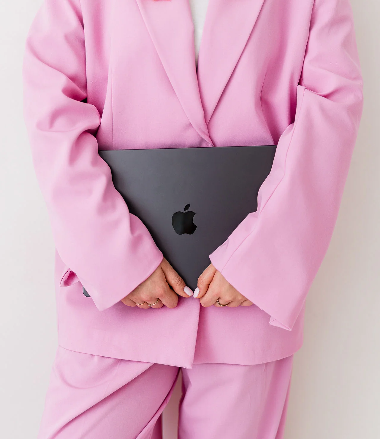 Kobieta w różowym garniturze stoi i trzyma w dłoni szary laptop.