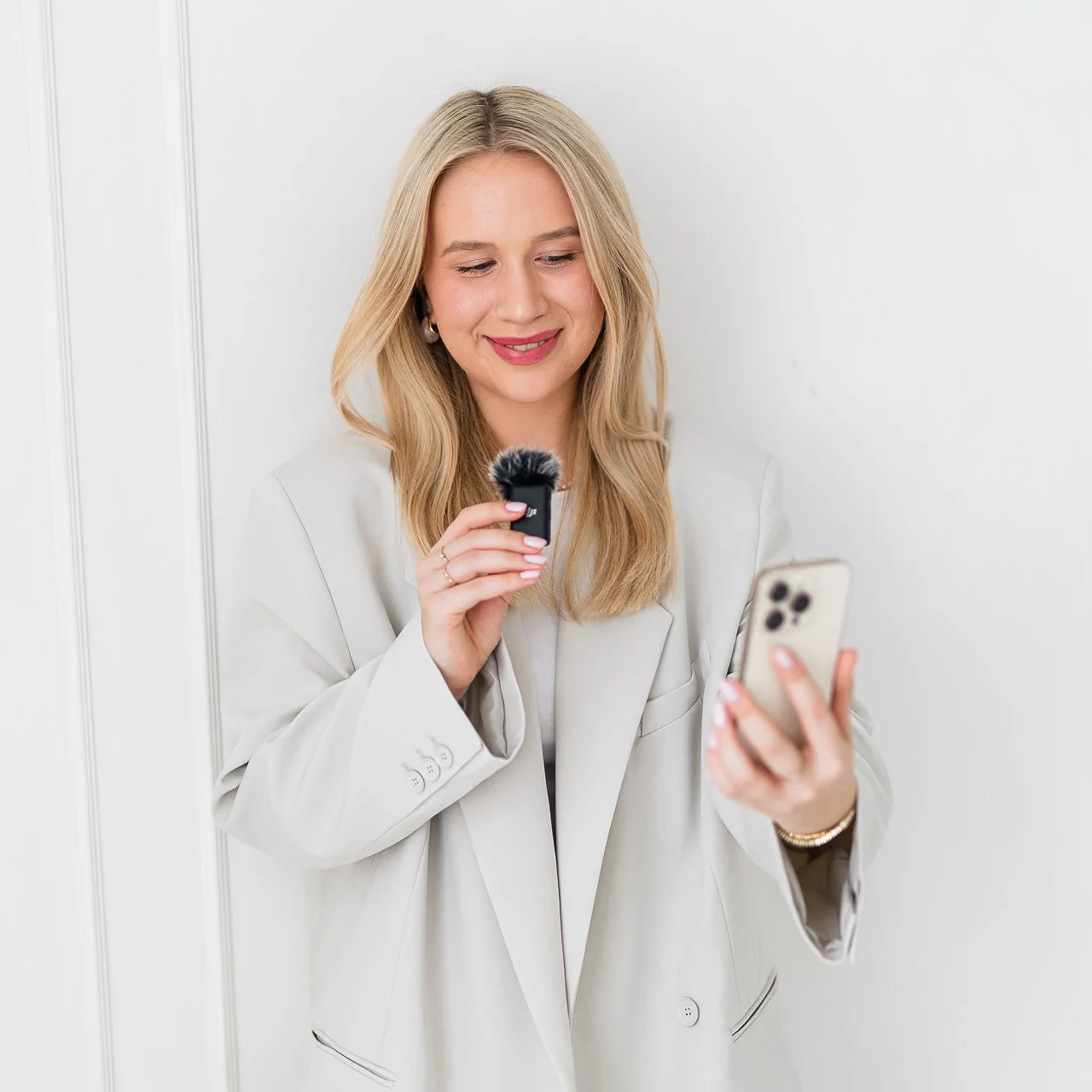 Młoda kobieta ubrana w stylowy biały garnitur w jednej dłoni trzyma mały mikrofon, a w drugiej telefon.